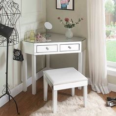 a white desk with two drawers and a stool in front of a window next to a flower vase