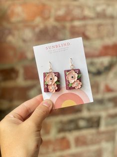 a person holding up a pair of earrings with flowers on them in front of a brick wall