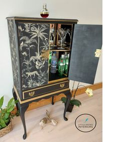 a black and gold painted cabinet with wine glasses on it's door, next to a potted plant