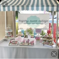 a table topped with lots of desserts under a striped awning next to a sign that says sugary sweet cottage