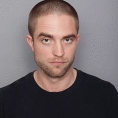 a man is looking at the camera while wearing a black t - shirt and standing in front of a gray wall