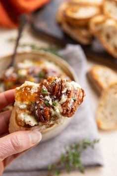 a hand holding up a piece of bread with cheese and nuts on it next to other food