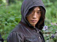 a young man wearing a hooded jacket in front of some trees and bushes, looking at the camera