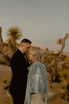 a man and woman standing next to each other in front of some cactus trees with their arms around each other