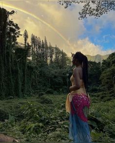 a woman standing in the middle of a forest with a rainbow in the sky behind her