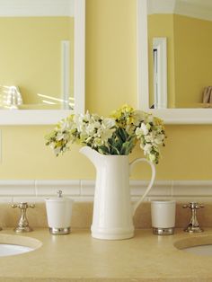 a white pitcher filled with flowers sitting on top of a bathroom counter next to two sinks