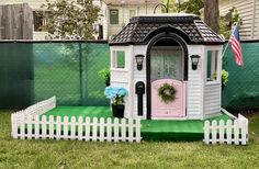 a small white house with a pink door and flower pot on the front lawn next to a picket fence