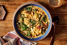 a blue bowl filled with pasta and spinach on top of a wooden table next to two spoons