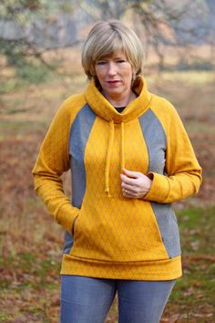 a woman wearing a yellow and grey hoodie standing in front of some trees with her hands on her hips