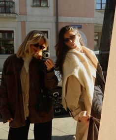 two women standing next to each other on the sidewalk with one holding a camera in her hand