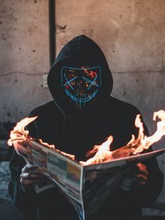 a person in a black hoodie with a neon mask on reading a news paper