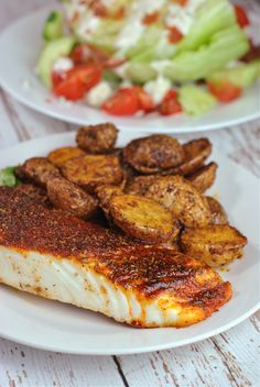 two white plates topped with fish, potatoes and salad on top of a wooden table