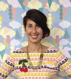 a woman wearing a colorful sweater and necklace with cherries on her chest, standing in front of a multicolored background