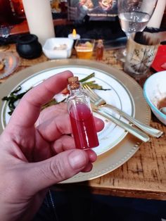 a person holding a small bottle filled with liquid on top of a wooden table next to plates