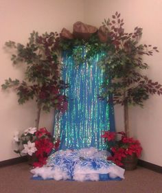 a room decorated for christmas with fake trees and flowers on the floor, in front of a blue curtain