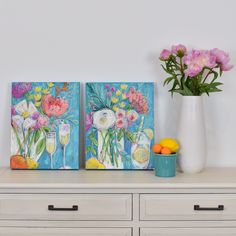 two paintings sitting on top of a dresser next to a vase filled with flowers and fruit