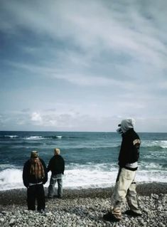 three people standing on the beach looking out at the water and rocks in front of them