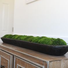 a black bowl filled with green moss sitting on top of a wooden dresser next to a white wall