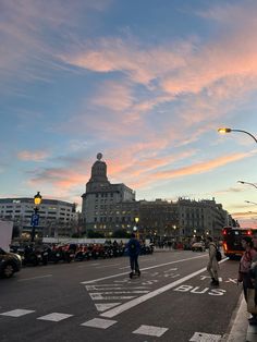 people are walking down the street in front of some buildings and cars at sunset or dawn