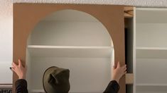 a person wearing a hat standing in front of a white book shelf with open doors