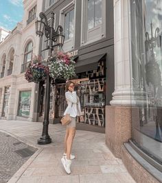 a woman standing on the sidewalk in front of a building with flowers hanging from it