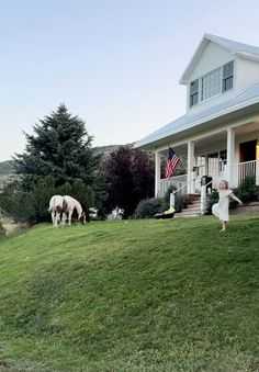 two horses graze on the grass in front of a white house with an american flag