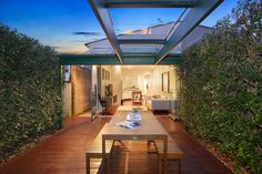 an outdoor dining area with wood flooring and wooden table surrounded by greenery at dusk