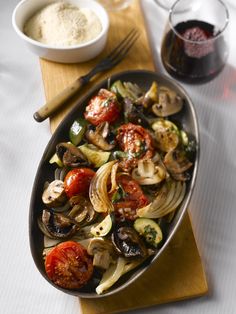 a pan filled with pasta and vegetables on top of a wooden cutting board next to a glass of wine