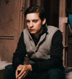 a young man sitting on top of a bed next to a wooden door and wearing a gray vest