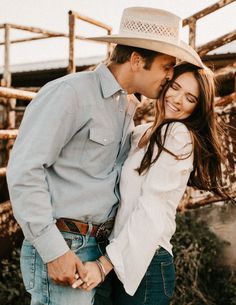 a man and woman standing next to each other in front of a wooden fence with hay