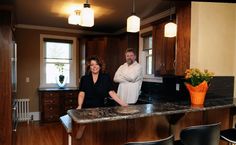 two people standing in a kitchen next to an island with granite counter tops and wooden cabinets