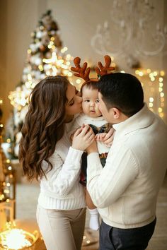 a young man and woman kissing their baby in front of a christmas tree with lights