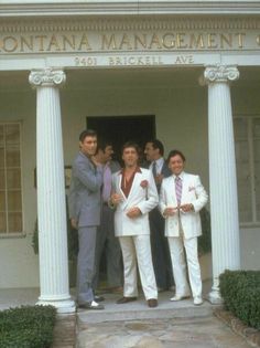 four men in white suits standing outside the front door of a building with columns and pillars