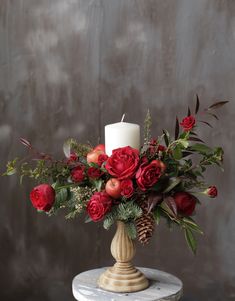 a white candle with red flowers and greenery on a marble pedestal in front of a gray wall