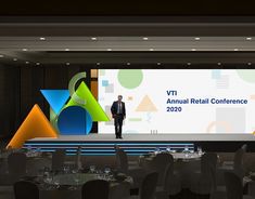 a man standing in front of a large screen at a conference hall with tables and chairs