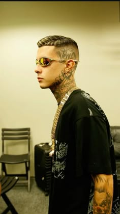 a man with tattoos on his face and neck standing in front of a chair wearing sunglasses