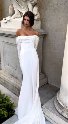 a woman standing in front of a statue wearing a long white dress with an off the shoulder neckline