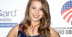 a woman with long brown hair and an american flag shirt smiling at the camera while standing in front of a white background