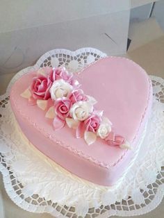 a pink heart shaped cake with white and pink flowers on the top is sitting on a doily