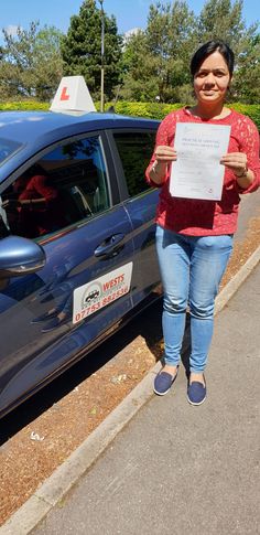 a woman standing next to a blue car holding up a piece of paper with writing on it