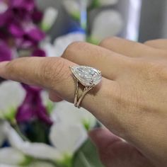 a woman's hand holding a ring with a pear shaped diamond on it and flowers in the background