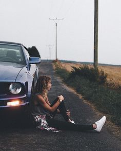 a woman sitting on the side of a road next to a blue mustang parked in front of her