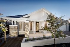 a white house with a black roof next to a tree and bushes in front of it