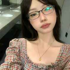 a young woman wearing glasses in a bathroom with sinks and mirrors behind her is looking at the camera