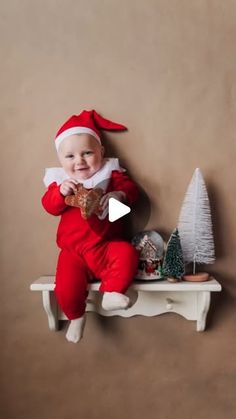 a baby dressed as santa claus sitting on a shelf next to a small christmas tree