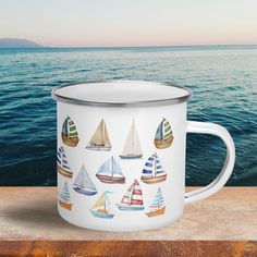 a white enamel mug with colorful sailboats on it sitting on a wooden table next to the ocean