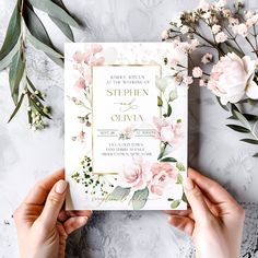 two hands holding up a wedding card with flowers and greenery around it on a table