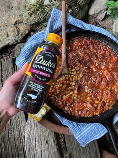 someone is holding a bottle of beer next to a skillet filled with baked beans