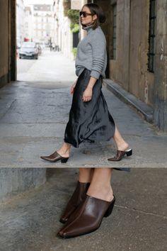 a woman in grey sweater and black skirt walking down the street with brown loafers