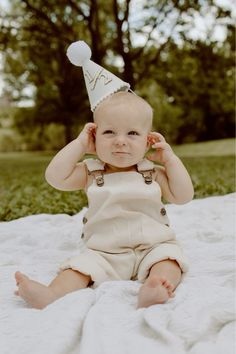 a baby wearing a party hat sitting on a blanket with his hands in the ears